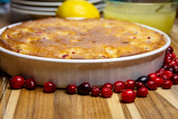 Fototapeta Freshly baked cranberry cake in a rustic dish, surrounded by scattered fresh cranberries and lemon. Homemade holiday dessert on dark wood.