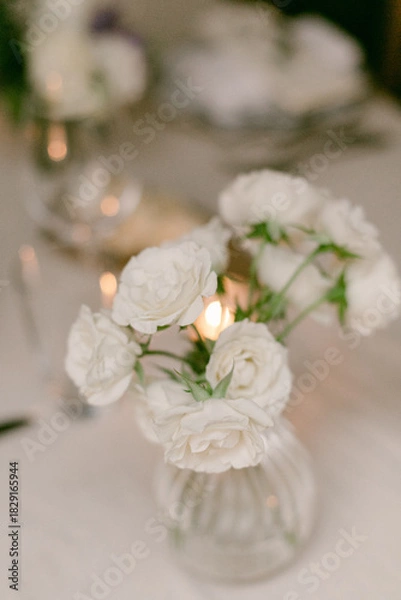 Fototapeta White roses in a small glass vase on an elegantly set dining table with warm candlelight