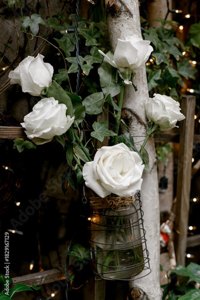 Fototapeta White roses in a glass jar hanging from a wooden structure surrounded by greenery and twinkling lights