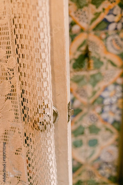 Fototapeta Delicate lace curtain partially drawn aside, revealing a colorful tiled wall in a cozy room