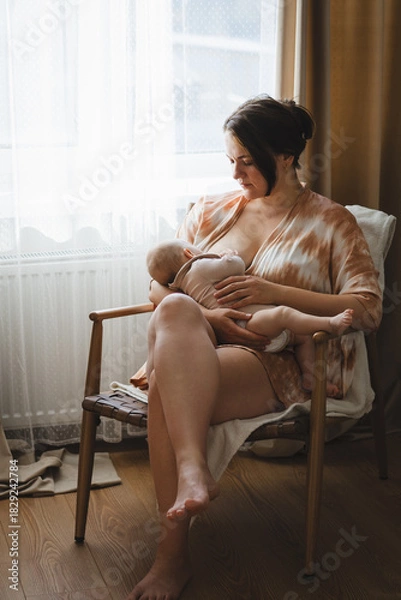Fototapeta A mother sits in a wooden chair, gently breastfeeding her baby in a warm and softly lit room. Sunlight filters through the curtains, creating a peaceful atmosphere.