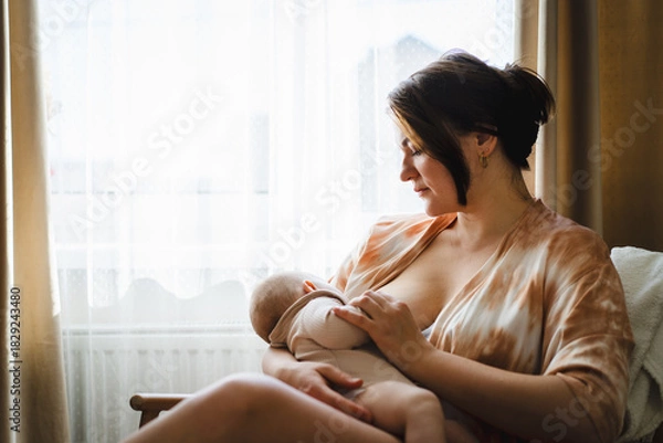 Obraz A mother sits in a wooden chair, gently breastfeeding her baby in a warm and softly lit room. Sunlight filters through the curtains, creating a peaceful atmosphere.