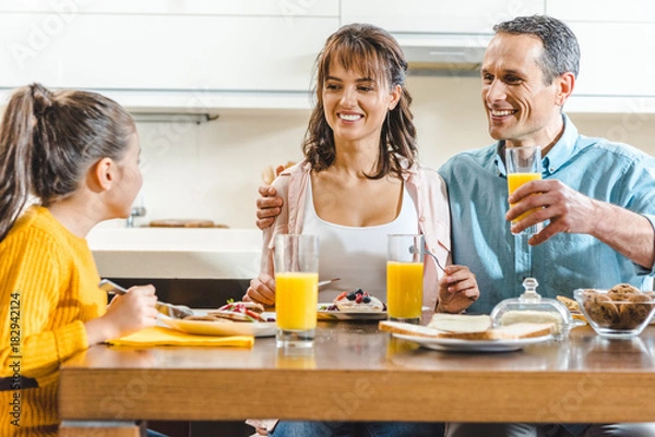 Fototapeta cheerful family sitting at table and holding glasses with juice at kitchen