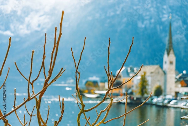 Obraz Bare winter branches frame the blurred scene of Hallstatt village, its church steeple, and the snow-dusted Alpine mountains rising dramatically across the dark lake.