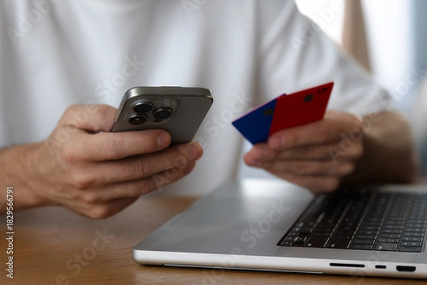 Fototapeta Man holds smartphone and credit cards next to laptop close-up