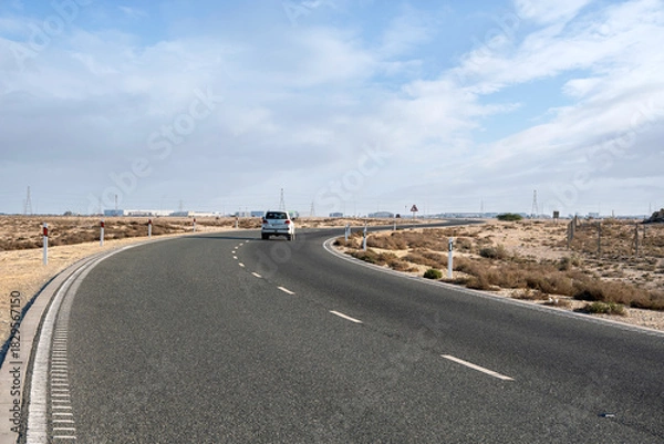 Fototapeta Dukhan, Qatar - November 25, 2025: A deserted asphalt road curving through a dry desert landscape. Road signs warn of an upcoming bend