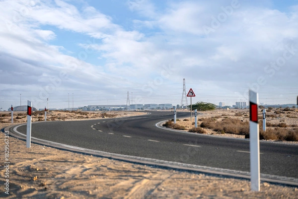 Fototapeta Dukhan, Qatar - November 25, 2025: A deserted asphalt road curving through a dry desert landscape. Road signs warn of an upcoming bend