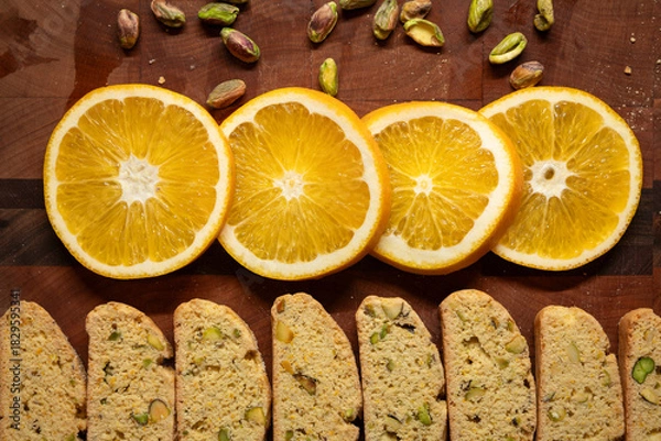Fototapeta Fresh orange slices, homemade biscotti cookies, and pistachio nuts arranged on a wood cutting board