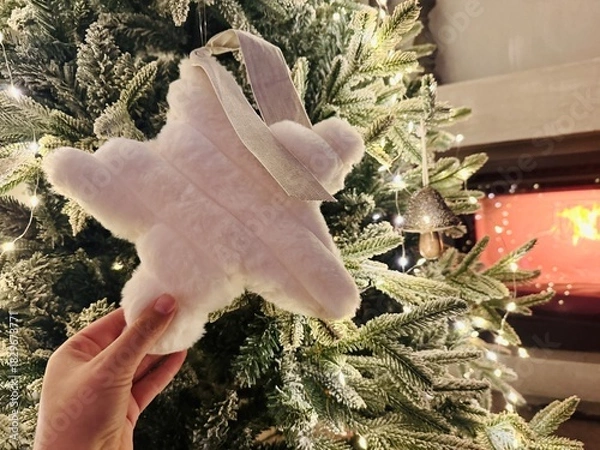 Fototapeta Hand Decorating Christmas Tree with Star Topper and Fireplace Burning Behind – Warm Festive Setting