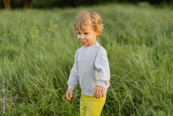 Obraz Playful toddler walking through tall grass, making a funny face in soft natural light. Candid outdoor moment showing real childhood personality and movement.