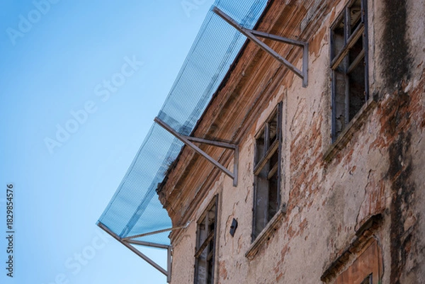 Obraz Old damaged building facade with broken windows and protective metal mesh installed above to prevent falling debris and ensure safety during planned renovation.