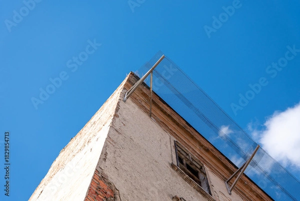 Obraz Old damaged building facade with broken windows and protective metal mesh installed above to prevent falling debris and ensure safety during planned renovation.