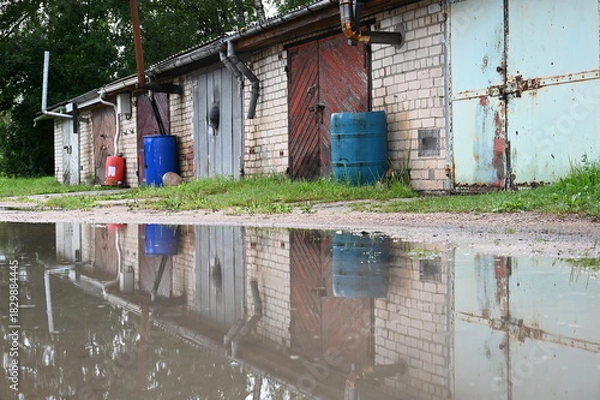 Fototapeta Lettland: Eine Reihe rustikaler Hinterhofgaragen spiegelt sich in einer Wasserpfütze