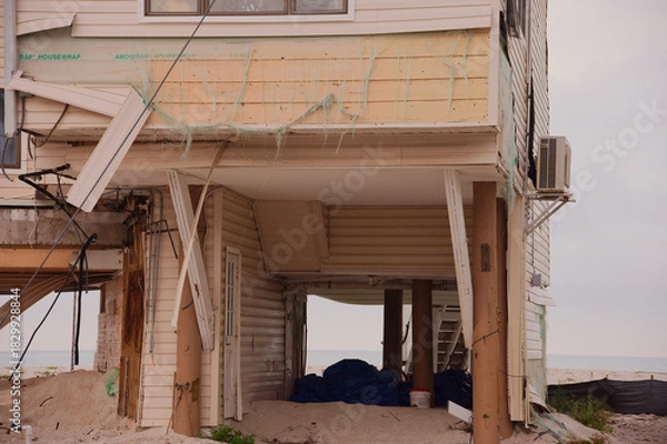 Obraz A partially elevated Beach House Damaged by Storm With Exposed Framework and Sand Underneath on the Shoreline in Treasure Island, FL. Sagging deck, and sand-filled ground. The scene captures coastal e