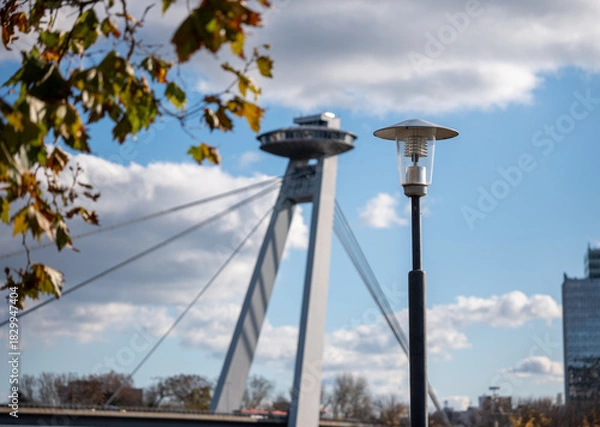 Fototapeta Street lamp in the foreground with the SNP Bridge and its UFO observation tower in Bratislava, Slovakia, captured on a sunny day with clouds and autumn leaves.