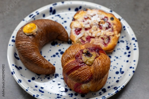 Obraz Plate with artisanal pastries including a banana-shaped banana cake, a fruity muesli cheesecake pastry, and a pistachio-filled pastry, perfect for breakfast or coffee break.