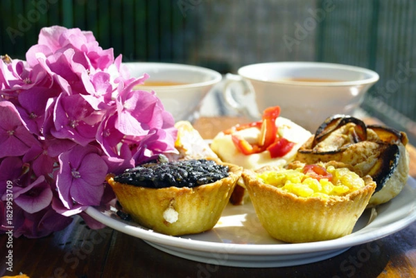 Obraz Tartlets filled with risotto and vegetables with pink hydrangea flowers beside tea cups on a rustic wooden table. Symbolizes afternoon tea elegance, garden party catering, and European bakery style.