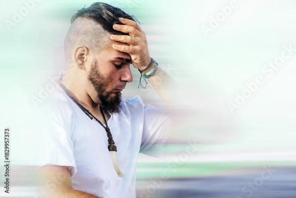 Fototapeta Young adult male with distressed expression holding forehead in motion-blurred background,