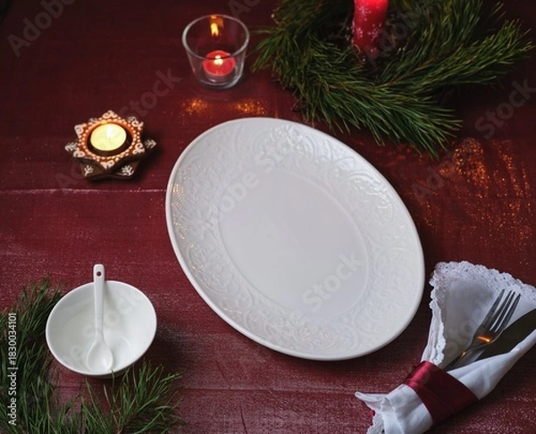 Fototapeta Christmas table setting on a burgundy tablecloth with a white oval platter, candles, and a napkin.