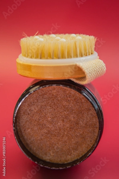 Fototapeta Natural body scrub jar with wooden lid and bristle brush on top, showcasing rich brown texture against a vibrant red background, emphasizing skincare and wellness