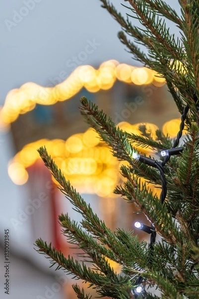 Fototapeta Close-up of a Christmas tree with string lights, with warm festive bokeh from a carousel or holiday lights in the blurred background