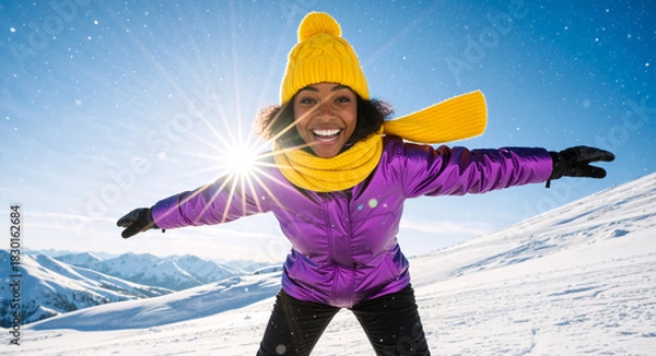 Obraz Happy young woman with open arms in snowy mountains. Cheerful female tourist in purple jacket and yellow hat enjoying winter vacation. Sunburst background