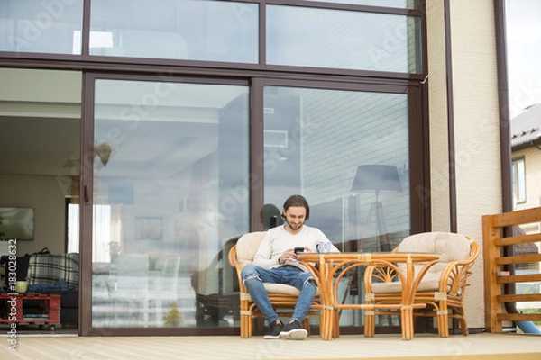 Fototapeta Young man sitting on chair at terrace of own modern house with glass wall, relaxing with cup of tea using mobile phone, attractive smiling guy enjoying spending time outdoors holding cellphone alone