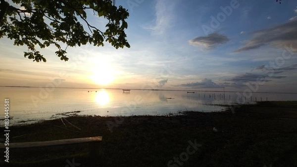 Obraz A beautiful sunset view over a vast expanse of water, with the shadows of trees and a bench on the lower left contrasting beautifully with the bright golden orange light and soft blue sky.