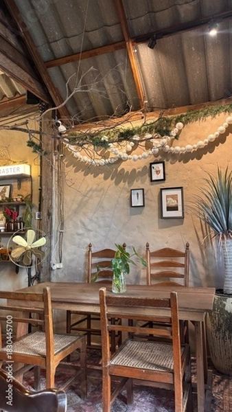 Obraz A cozy, retro-inspired lounge area in a coffee shop features a set of four wooden tables and chairs, earth-colored walls decorated with hanging pictures, plants, antique fan lamps