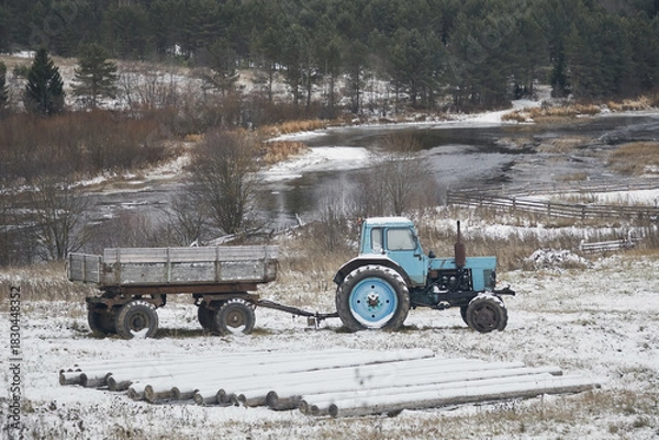 Fototapeta An old tractor with a cart in winter in a field with snow and logs near the river