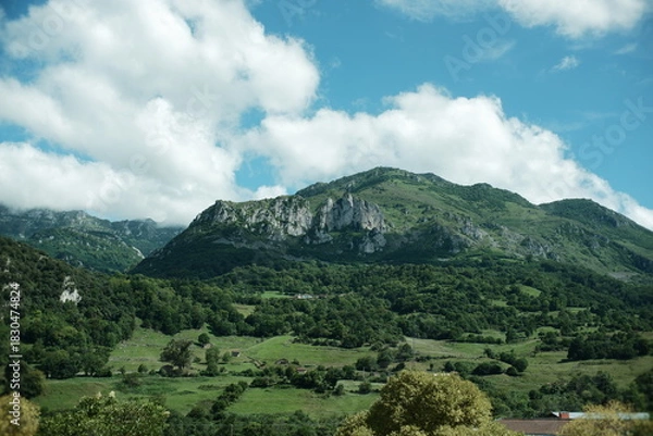 Fototapeta Abstract landscape in northern Spain, with green meadows and trees in the foreground and partially vegetated mountains under a blue sky with clouds. Rural peaceful landscape