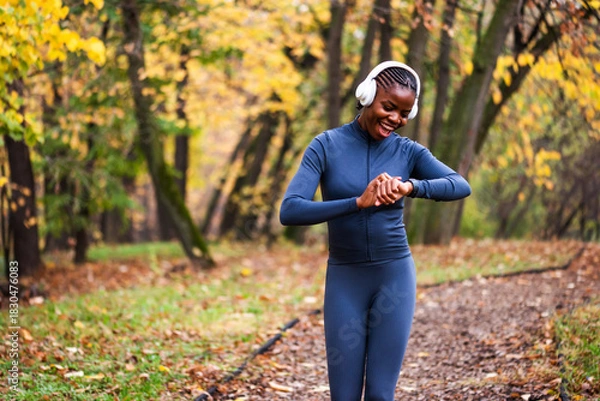 Fototapeta A runner in dark athletic wear moves confidently down a wooded path, framed by vibrant fall foliage and soft natural light. She is looking at smartwatch.