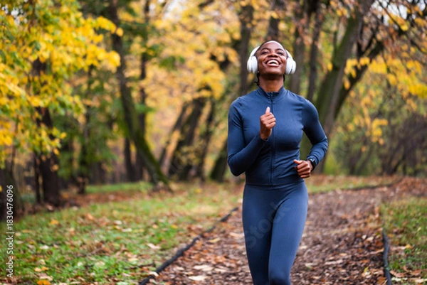 Fototapeta Happy woman jogs along a leaf-covered forest trail, surrounded by warm autumn colors and tall trees as she enjoys an outdoor run.