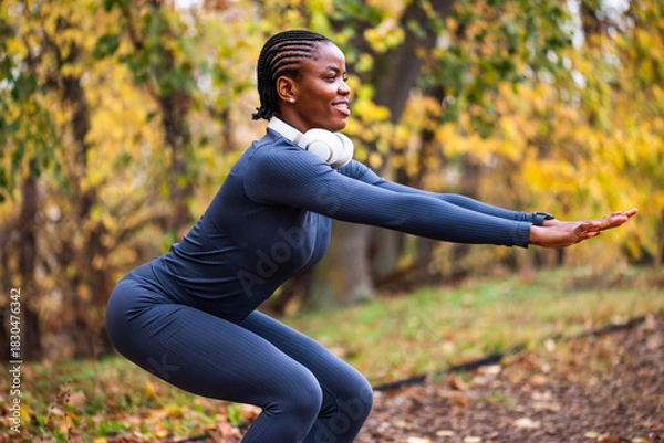 Fototapeta Young black woman is exercising in park in autumn. She is doing squats.