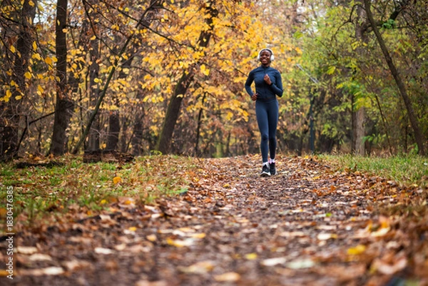 Fototapeta A smiling black woman runs on running trail of fallen leaves, with golden and green trees creating a serene, seasonal backdrop.