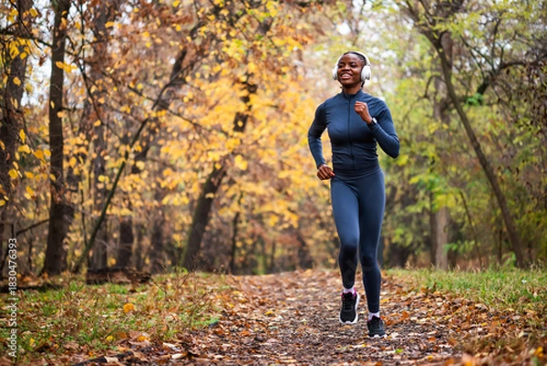 Fototapeta A smiling black woman runs on running trail of fallen leaves, with golden and green trees creating a serene, seasonal backdrop.