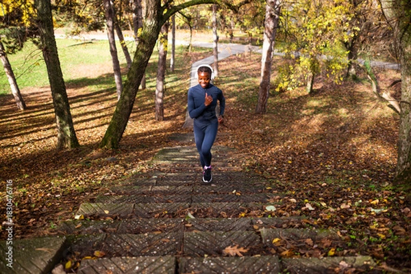 Fototapeta A woman jogs up a leaf-covered stone path in a park, wearing a navy athletic outfit and headphones, enjoying a sunny autumn day.