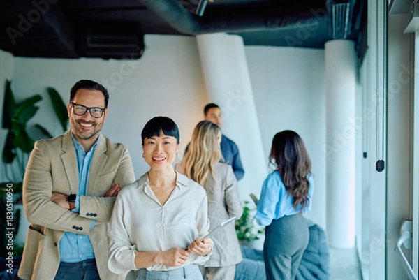 Obraz Diverse business professionals smiling, looking at camera in a bright, contemporary office. Coworkers collaborating in the background