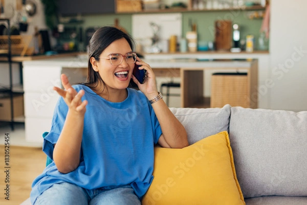 Fototapeta The excited young woman energetically chats on her phone, surrounded by a beautifully decorated living space, reflecting her vibrant personality and engagement.