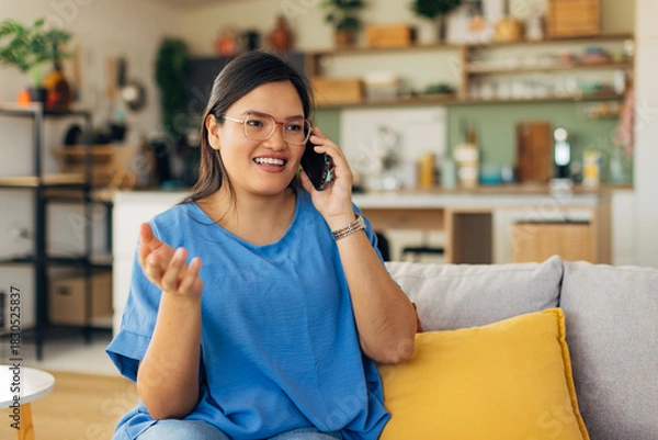 Fototapeta A joyful young woman is seen smiling and gesturing while talking on her phone in a cozy living room, reflecting a modern and vibrant lifestyle filled with positivity.