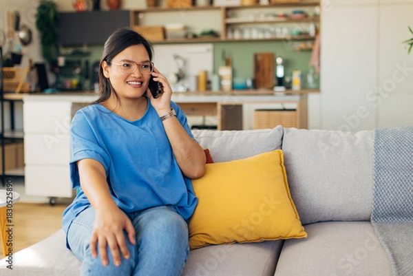 Fototapeta Seated comfortably in her stylish living room, a young woman enjoys a phone chat, showcasing emotional engagement and the warmth of domestic life amidst a carefully curated space.