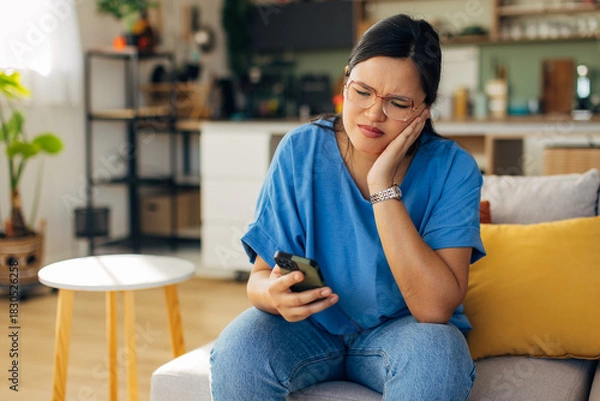 Fototapeta A young woman appears stressed while looking at her smartphone, portraying the impact of digital communication on personal emotions in a cozy home environment.