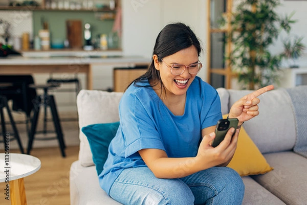 Fototapeta A young woman joyfully engaging with someone through her smartphone during a video call while seated comfortably in a cozy living room space full of greenery.