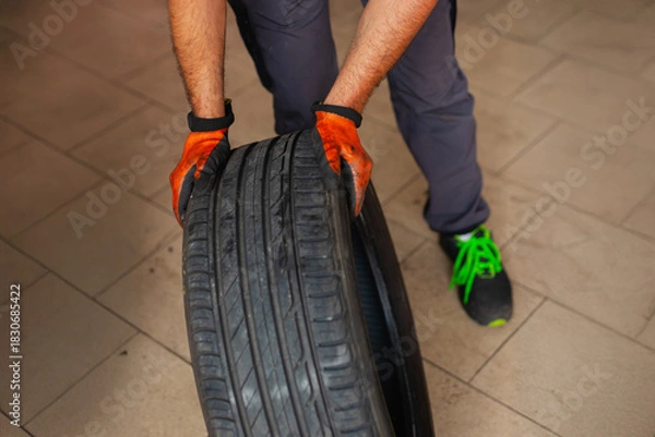 Fototapeta Mechanic with orange gloves moving a tire in the workshop. Car maintenance and tire change for road safety.