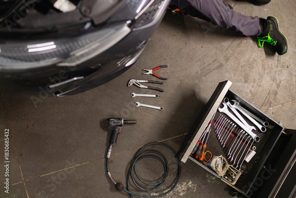 Fototapeta Mechanic under car with tools on floor: wrenches, pliers, impact gun and tool chest. Auto repair in professional workshop.