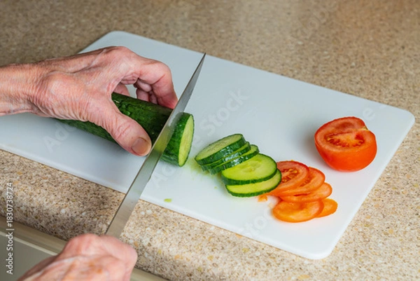Fototapeta Older person preparing salad items with a cucumber and tomato on a white board