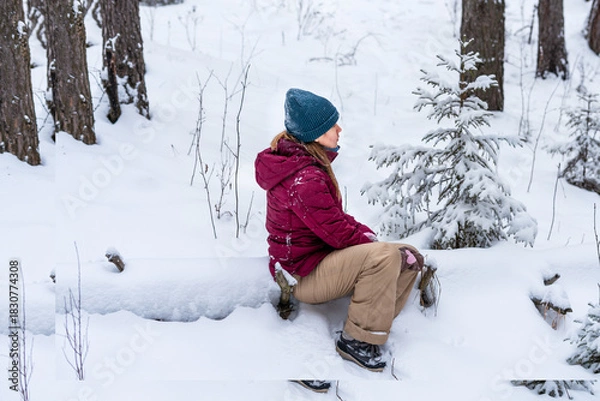 Fototapeta A young Caucasian woman sits on a snow-covered log in a winter forest and resting She wears a maroon jacket and a blue beanie, surrounded by snow and evergreen trees