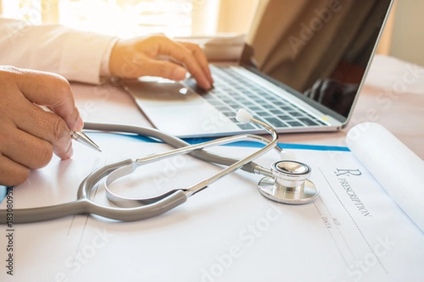 Fototapeta Doctor's writing and working on laptop computer, writing prescription clipboard with record information paper folders on desk in hospital or clinic, Healthcare and medical concept. Focus stethoscope