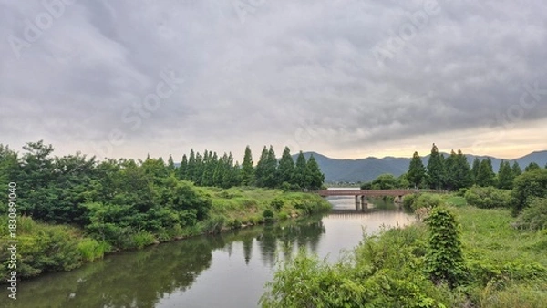 Fototapeta Lake surrounded by lush green trees and distant hills under soft cloudy daylight, forming a peaceful natural scenery.