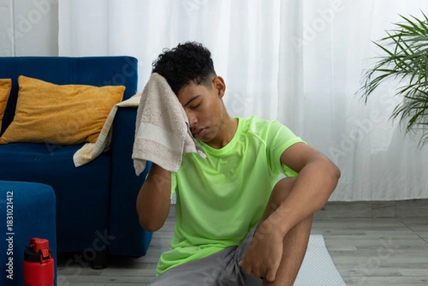Obraz Brown skinned teenager sitting on an exercise mat in his living room, wiping off sweat after working out. Image of effort, well being, and home training